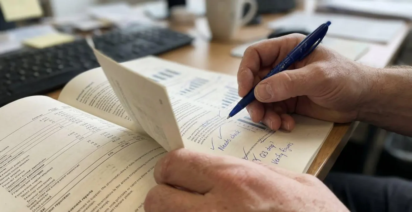 Gros plan sur des mains feuilletant un rapport annuel avec des annotations au stylo, arrière-plan de bureau flouté